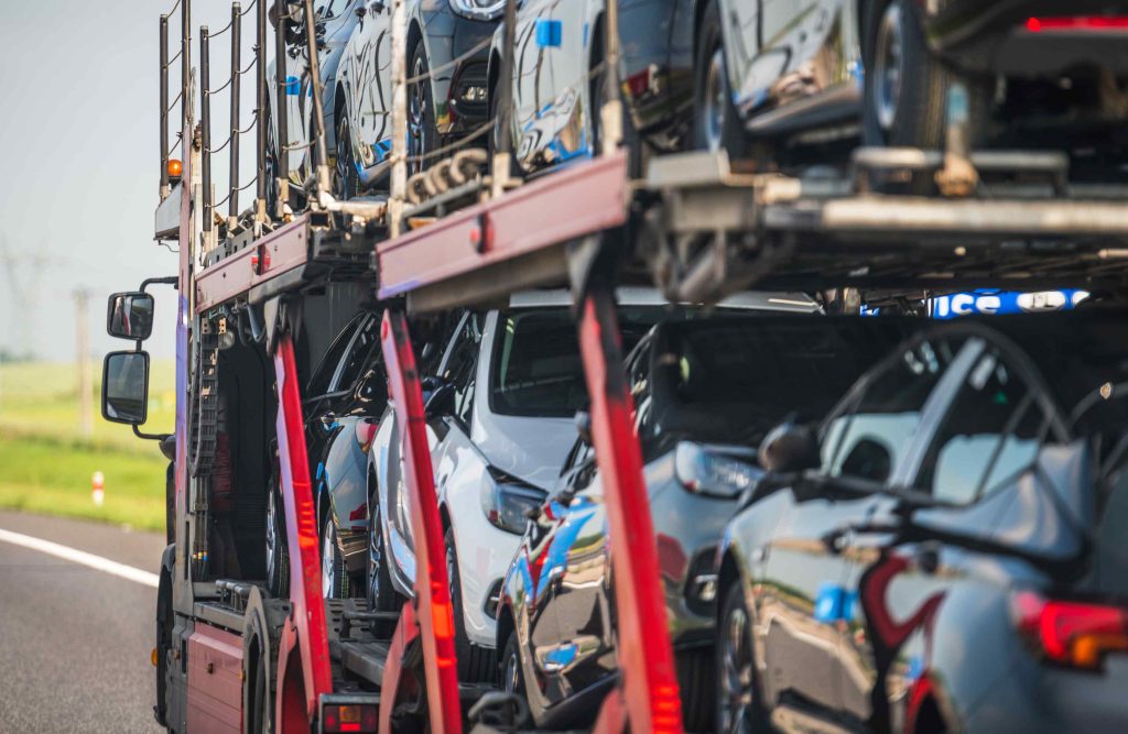 A car carrier truck, part of a Car Transport Tallahassee, FL service, transports multiple new cars on a highway. The truck is loaded with vehicles on both its upper and lower decks. The scene takes place on a sunny day with roadside grass visible.