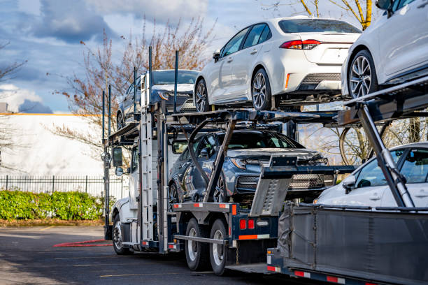 A car carrier truck loaded with new vehicles is parked outdoors on a sunny day, with several white and silver cars visible on the trailer—an everyday scene in FL Car Shipping Tallahassee. Trees and a fence are in the background, highlighting the reliable FL auto transport services available in Tallahassee.