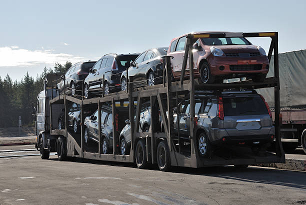 A car carrier truck loaded with two levels of various cars is parked outdoors, with trees and another vehicle visible in the background under a clear sky—showcasing a typical scene for FL Car Shipping Tallahassee.