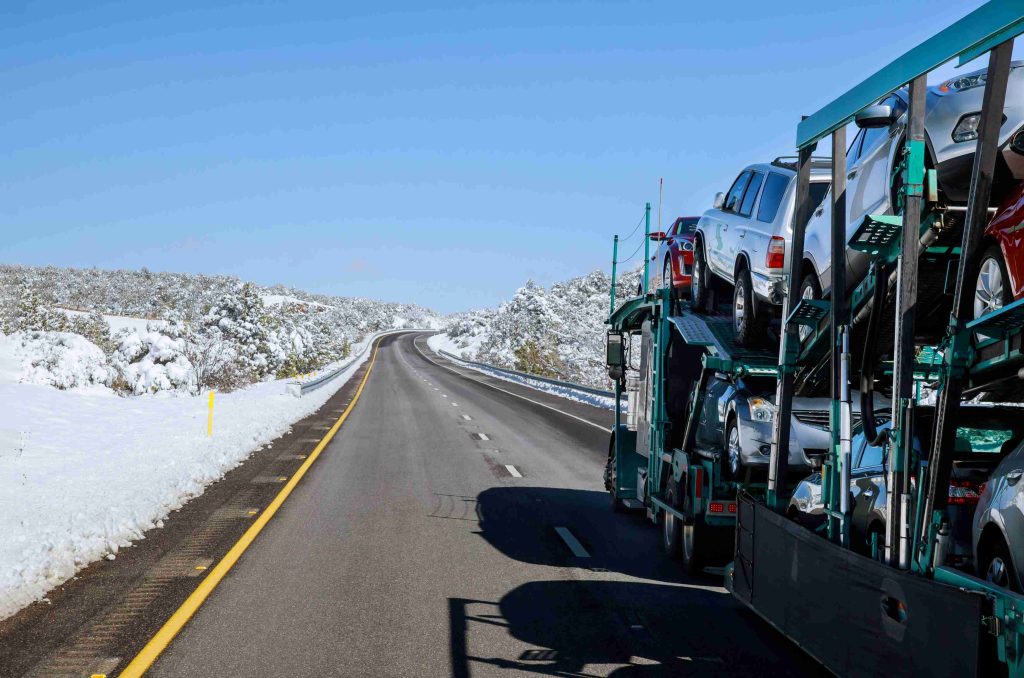 A car carrier truck loaded with vehicles drives on a highway surrounded by snow-covered landscape under a clear blue sky, illustrating the reliability of Auto Transport Tallahassee services even in challenging winter conditions.